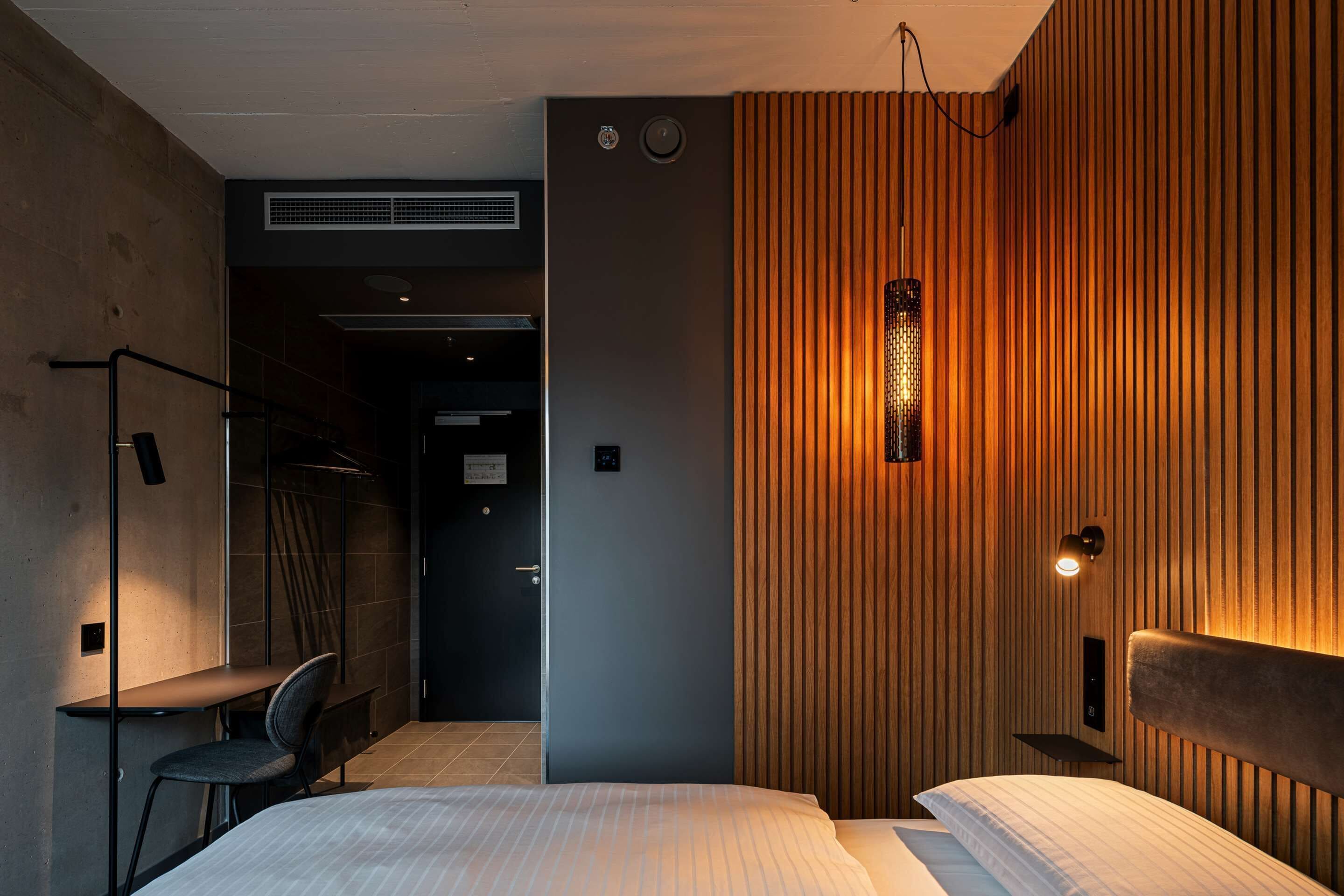 A modern hotel room with a bed, a desk, and a chair. There is a wooden panel wall with a light fixture.