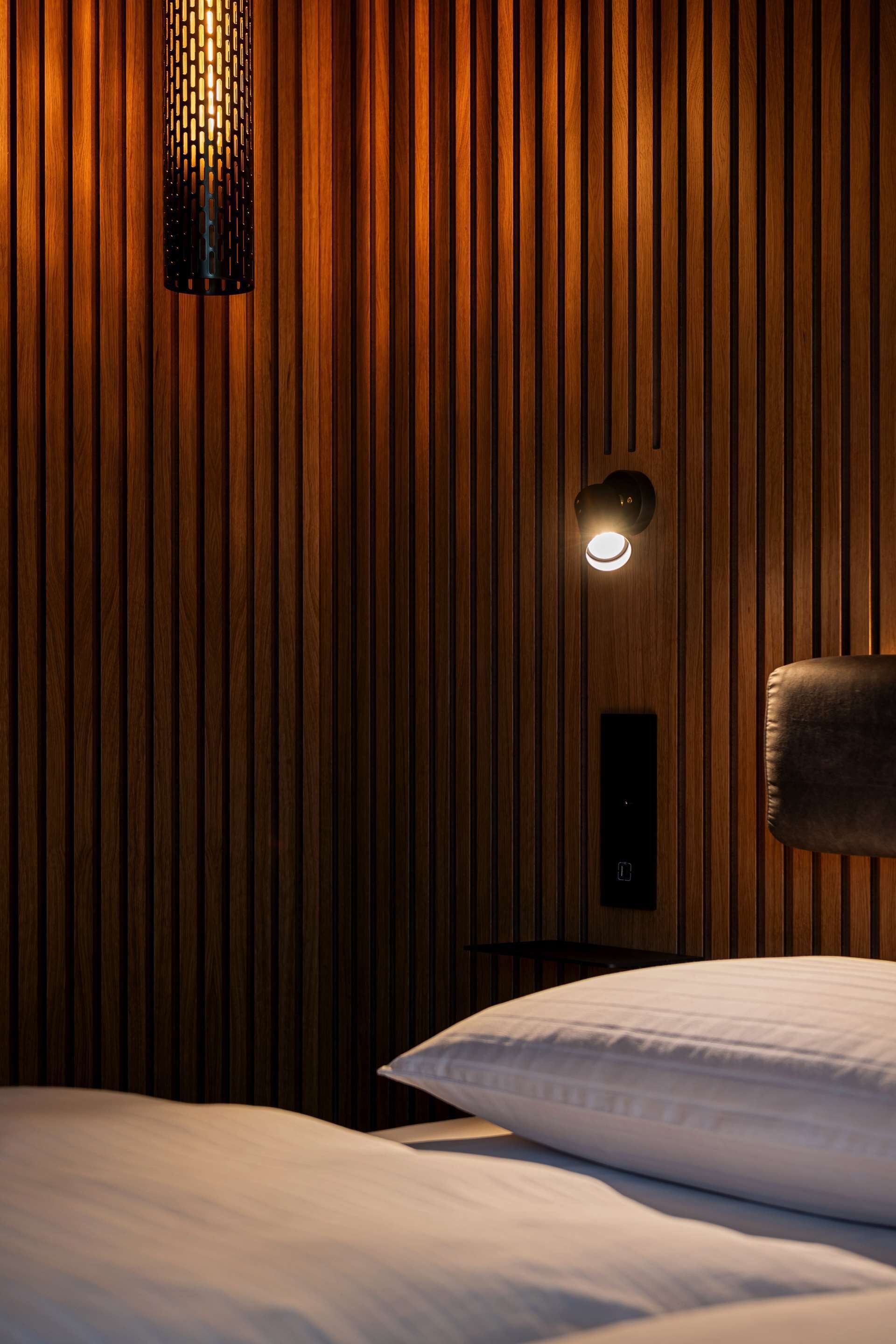 A hotel room with a wooden panel wall, a bed with white bedding, and a wall-mounted light fixture.