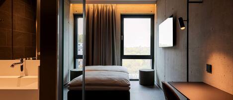 A modern bedroom with a large bed, a desk, a chair, a TV, and a view of the outdoors through a large window.