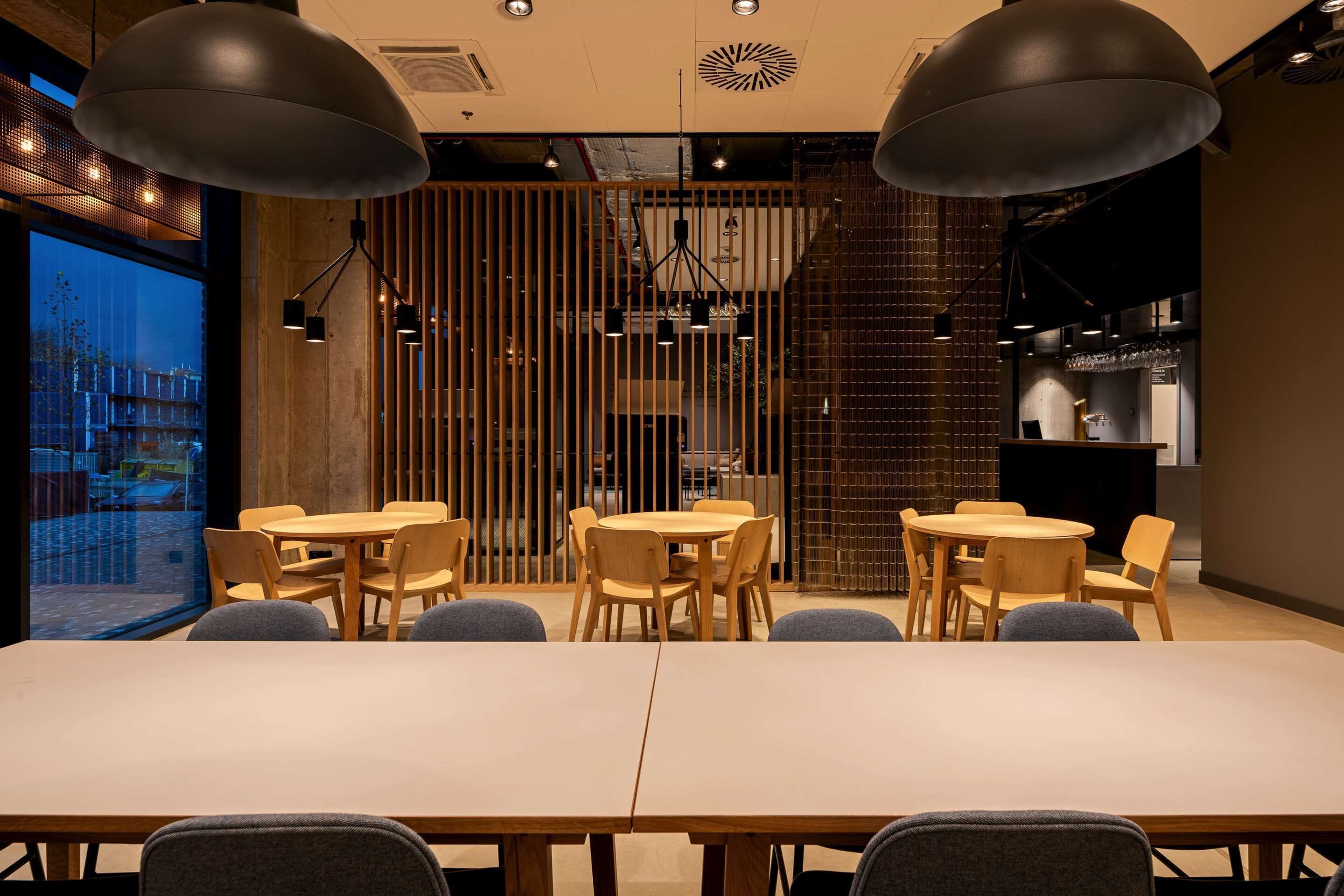 Un espace de restauration moderne avec des tables et des chaises en bois, des luminaires suspendus et une vue dégagée sur l’extérieur grâce à une grande fenêtre.