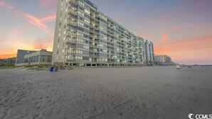 Un hotel de varios pisos con fachada plana, situado en una playa arenosa con un atardecer de fondo.