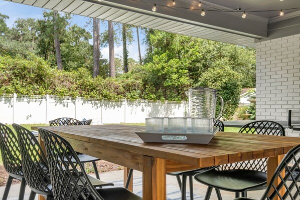 Un espacio de comedor al aire libre con una mesa y sillas de madera, una jarra de vidrio transparente y una pared de ladrillo blanco.