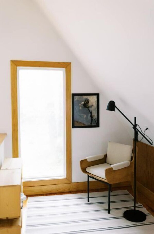 Um quarto com teto inclinado, janela com moldura de madeira, uma cadeira branca, abajur de chão preto e um quadro na parede.