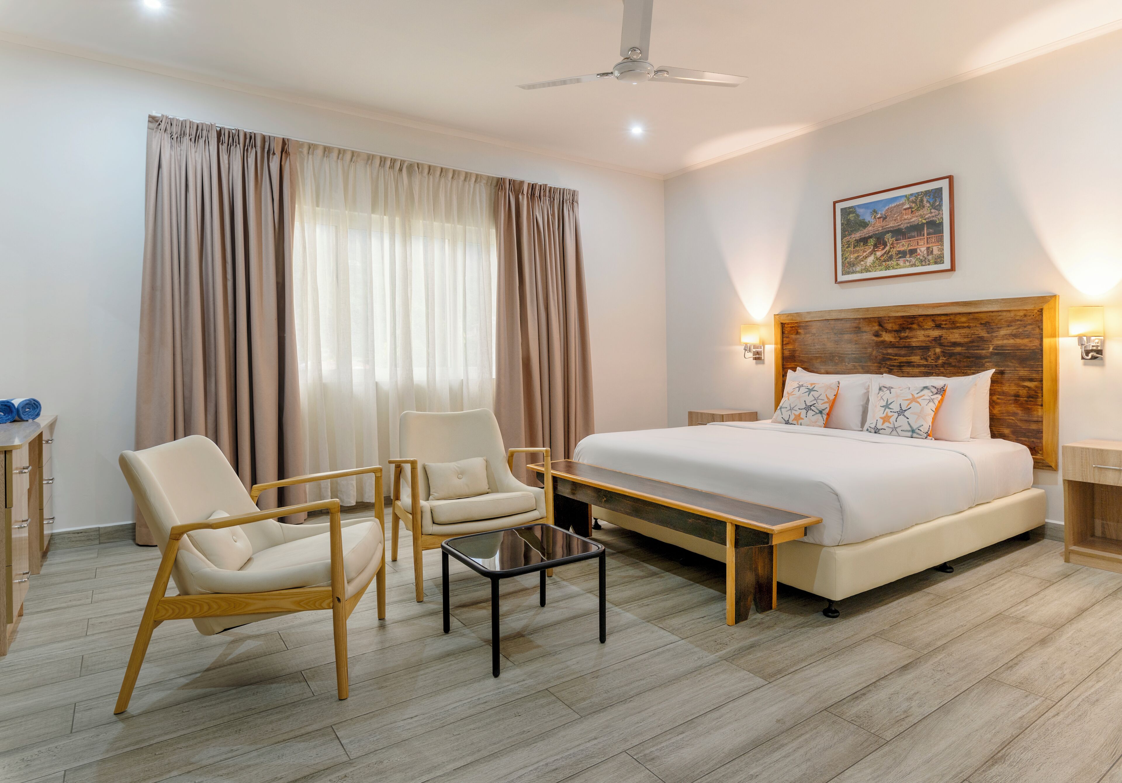 A hotel room with a bed, two chairs, a small table, a ceiling fan, and a painting on the wall.