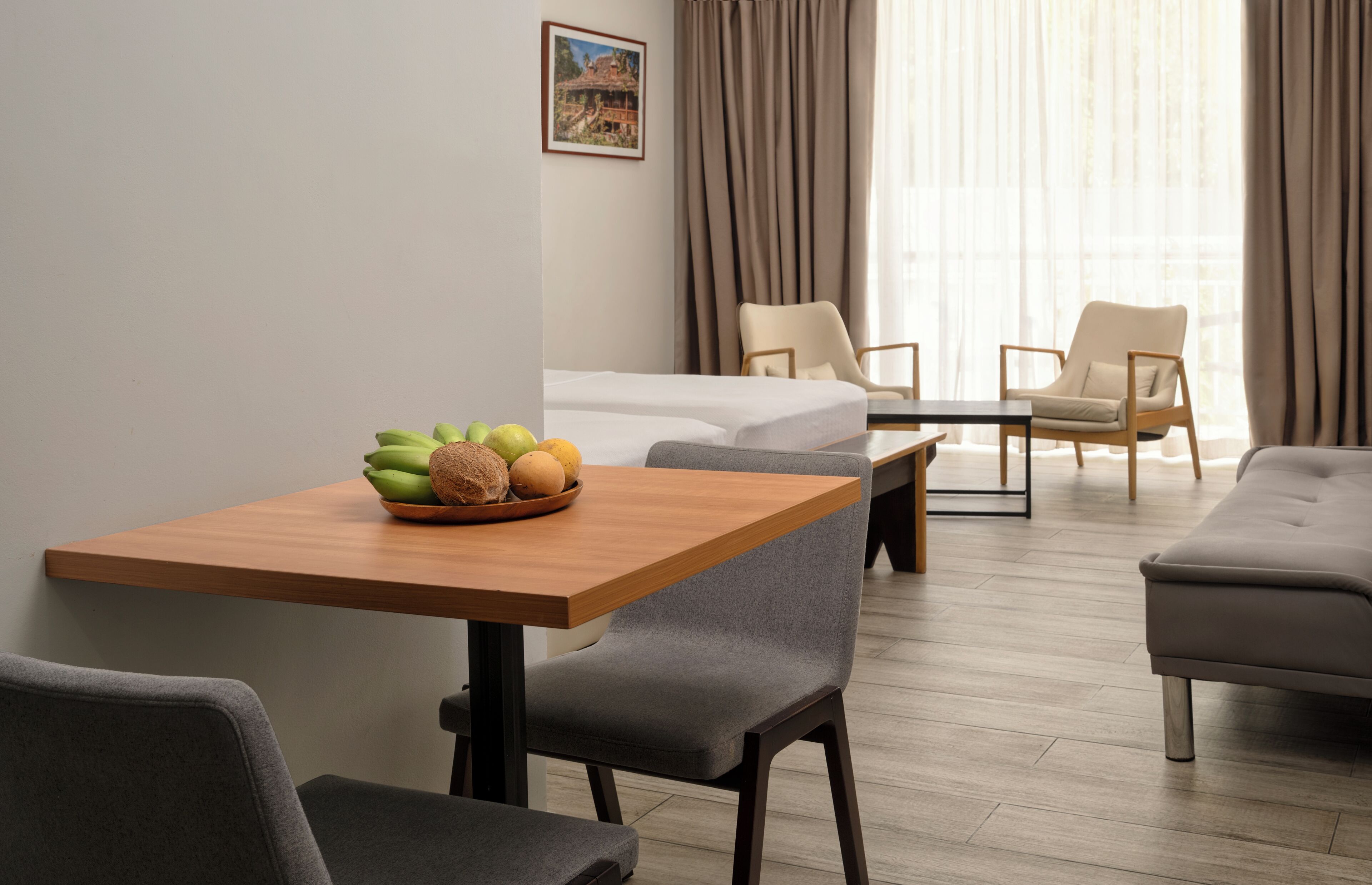 Una habitación de hotel con una mesa de madera, dos sillas grises, una cama y un ventanal con cortinas.