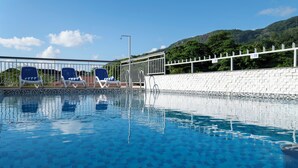 Ein Swimmingpool mit klarem, blauem Wasser, umgeben von einer weißen Mauer und einem Metallgeländer. Liegestühle säumen den Poolrand.