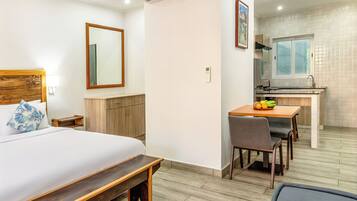 A hotel room with a bed, bedside table, wooden headboard, a small desk with a chair, a window, and a kitchenette.