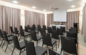 A conference room with rows of chairs, a projector screen, a table with a white tablecloth, and curtains. - Voilà Beau Vallon (Mahé Island)