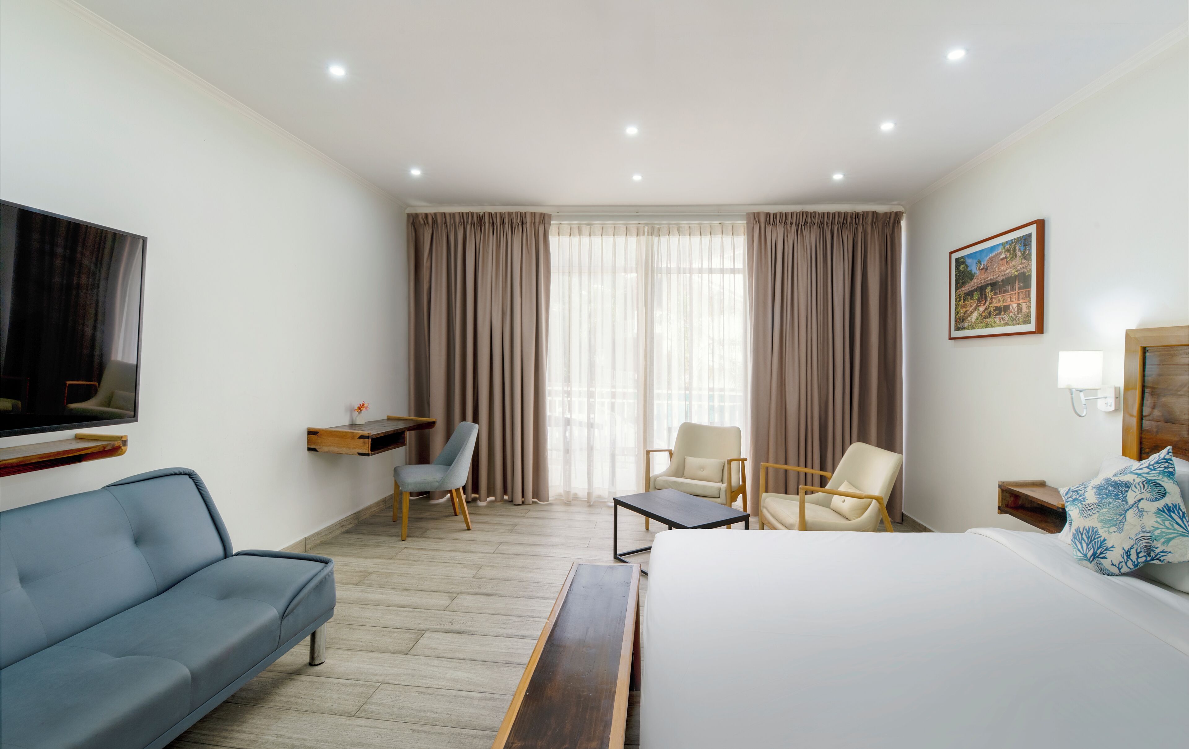 Une chambre d’hôtel avec un lit, un canapé, deux fauteuils, un bureau, une télévision et un tableau au mur.
