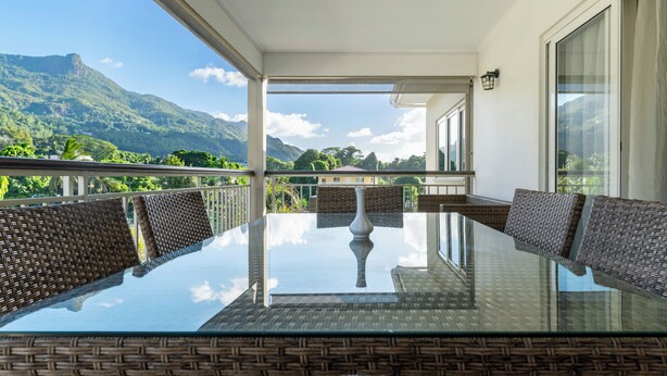 Ein Balkon mit Glastisch und Korbstühlen, der auf eine gebirgige Landschaft blickt.