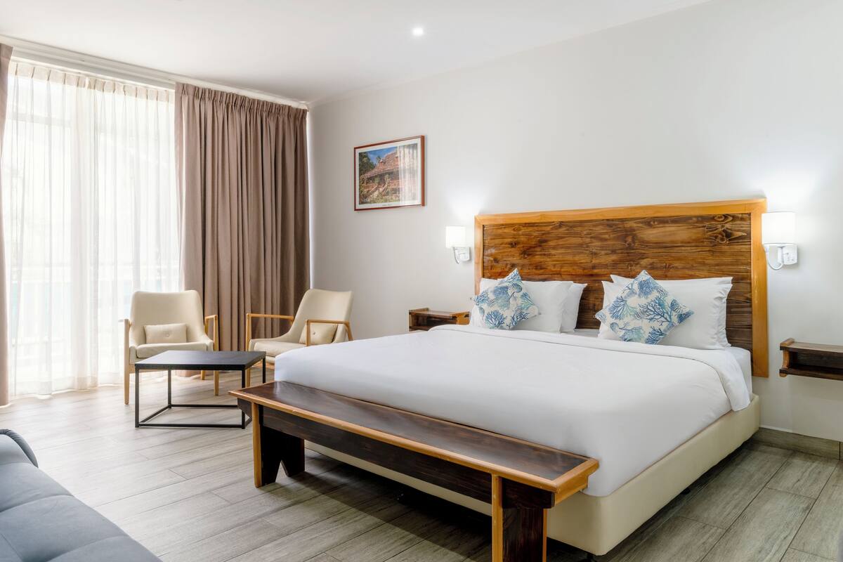 A hotel room with a large bed, two chairs, a bench, a wooden headboard, a nightstand, and a painting on the wall.