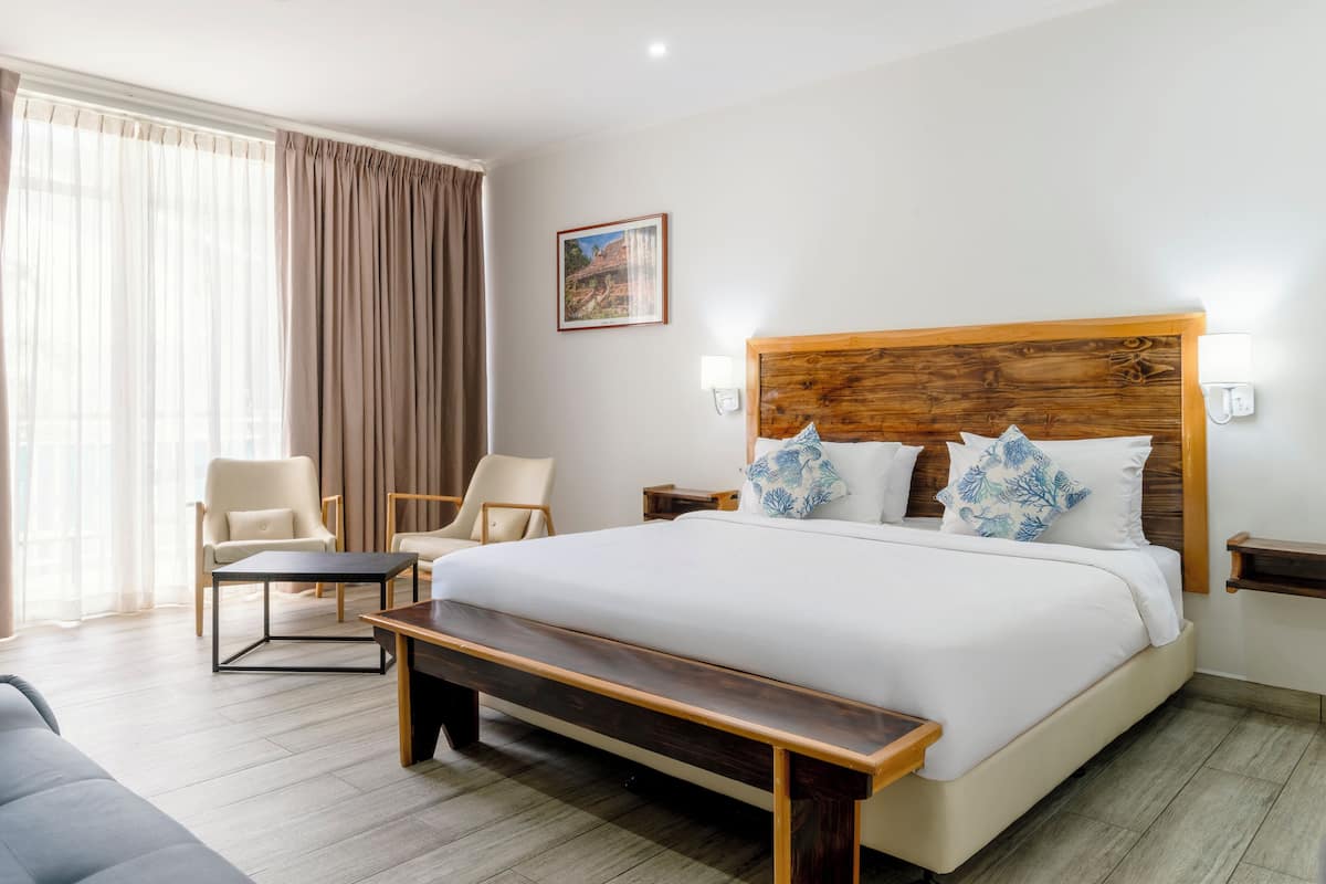 A hotel room with a large bed, two chairs, a bench, a wooden headboard, a nightstand, and a painting on the wall.