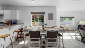 A modern kitchen with white cabinets, a dining area with a table and chairs, and large windows offering a view of a white picket fence and greenery. - The Gather House! Super clean, Super comfortable and Super private! (The Dalles)
