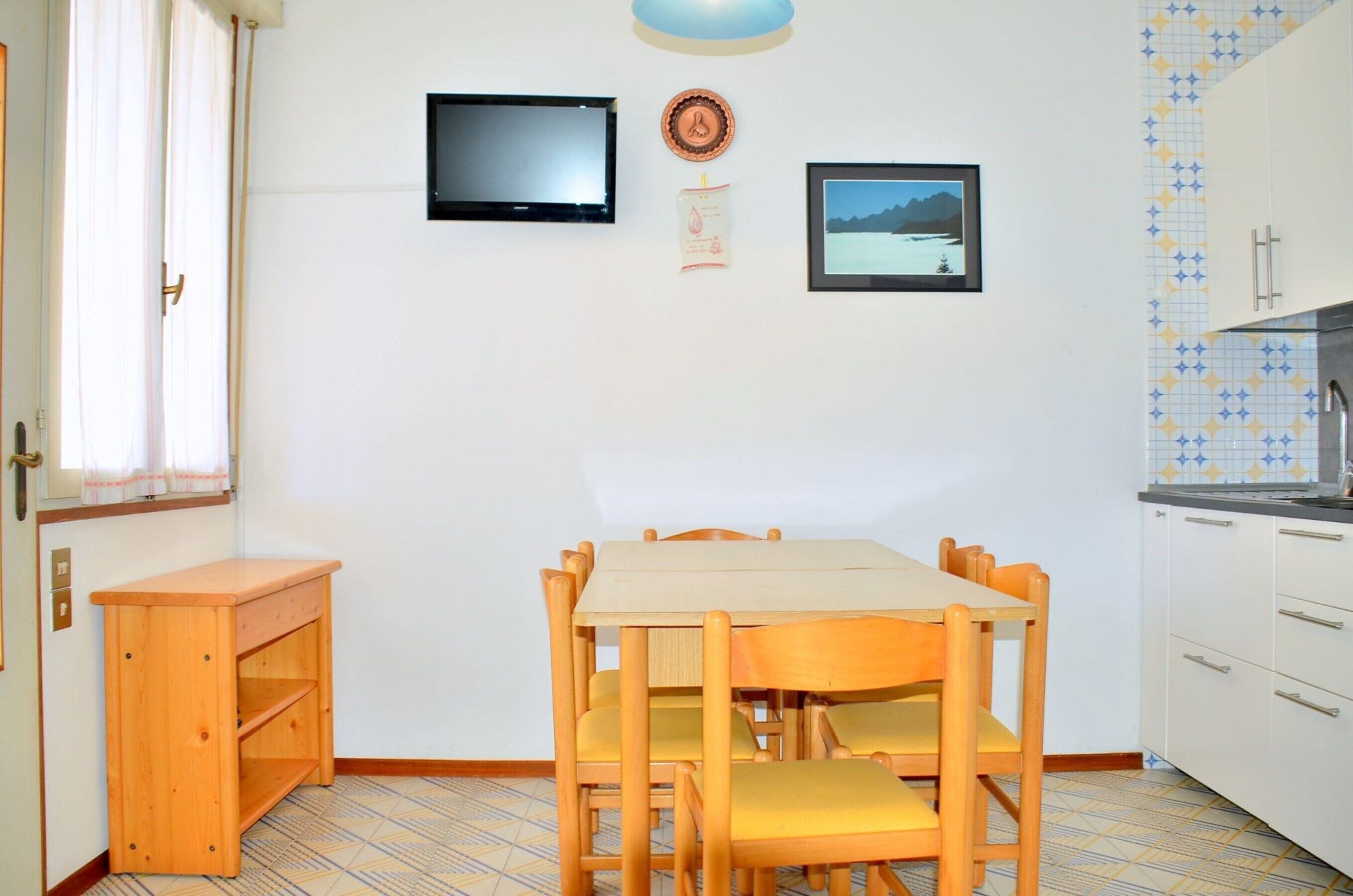 A compact kitchen with a dining table and chairs, a small wooden cabinet, and a television mounted on the wall.