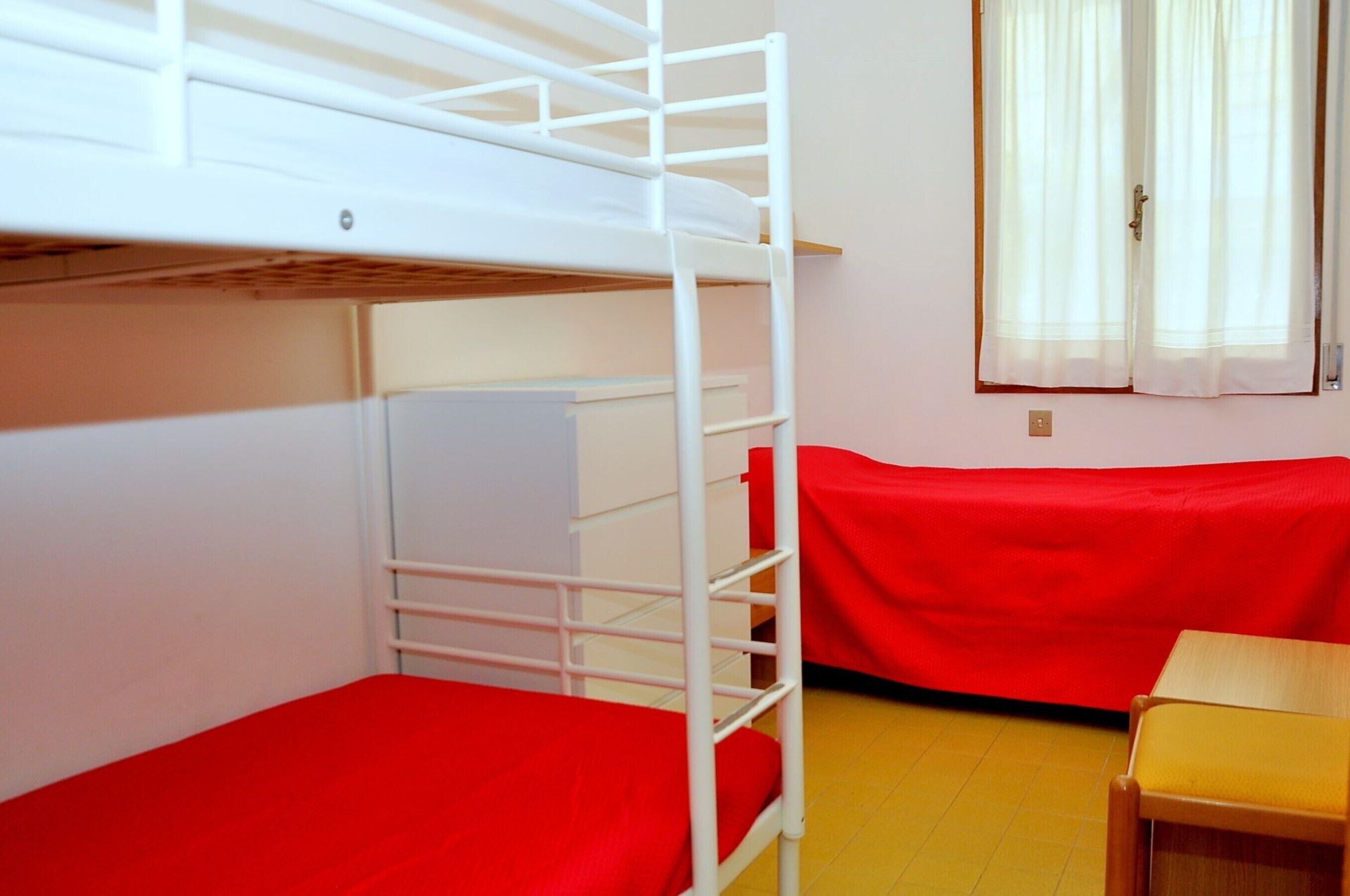 Quarto com beliche, colchão vermelho, mesa de madeira e janela com cortinas.