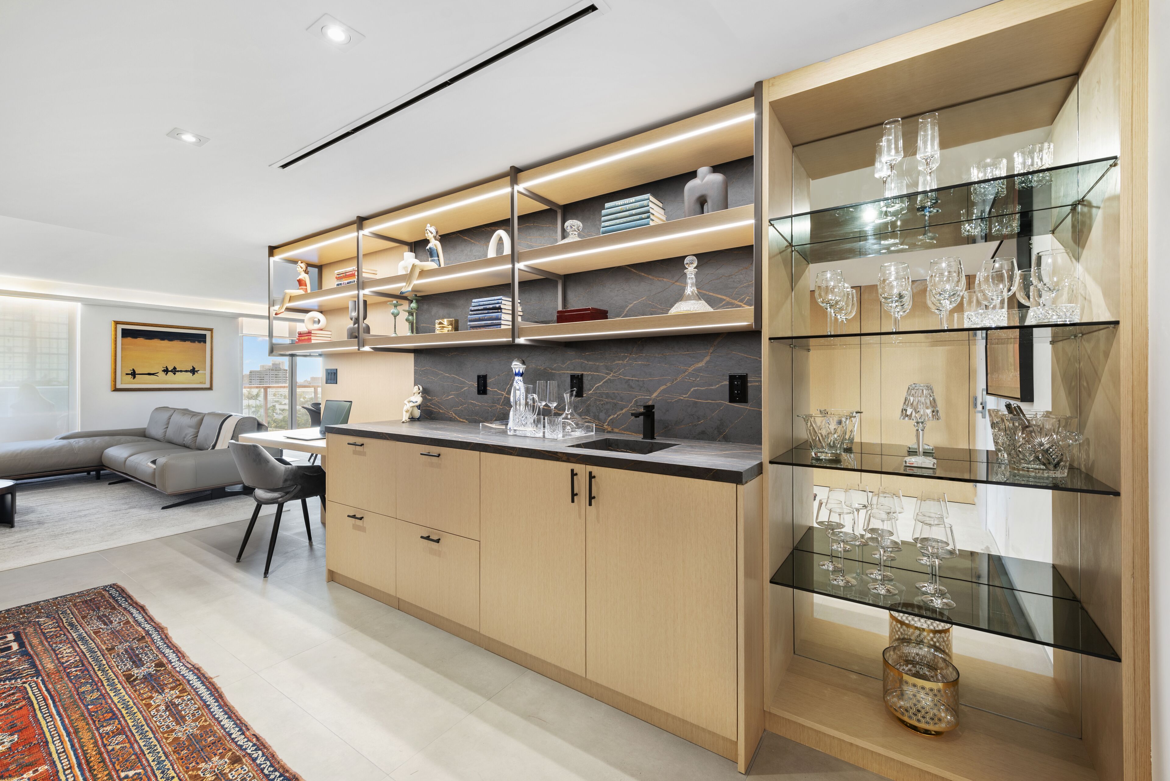 A modern kitchen with built-in shelves displaying glassware and decorative items, a dining area with a sofa and chairs, and a patterned rug on the floor.