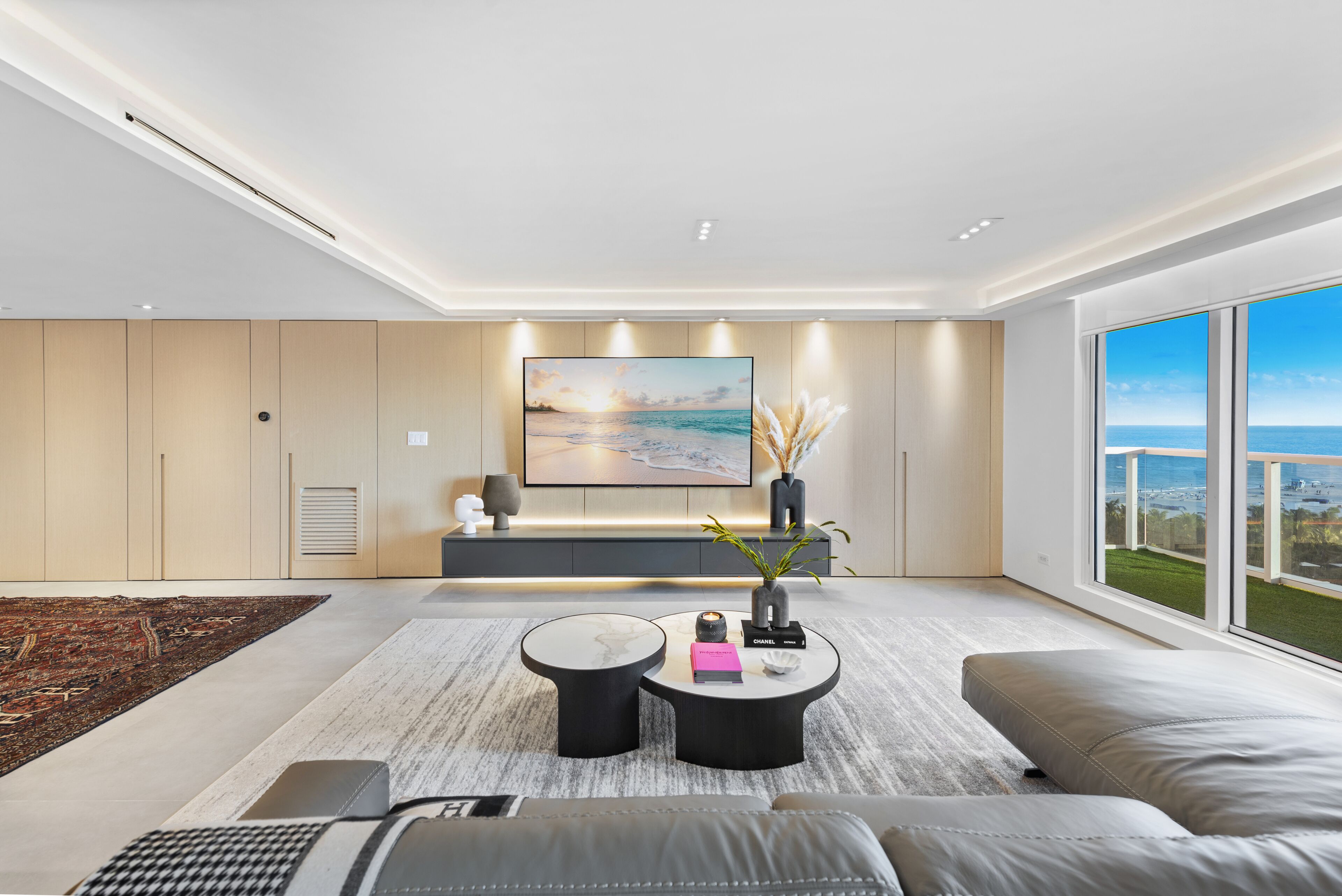 A modern living room with a large flat-screen TV, a sofa, two coffee tables, and a view of the ocean through large windows.