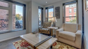 Una sala de estar moderna con ventanas amplias, un sillón estampado, un sofá beige y una pequeña mesa con una lámpara.