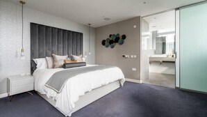 Une chambre à coucher moderne avec un grand lit, une tête de lit grise, une table de chevet, un luminaire fixé au mur et un élément décoratif mural.