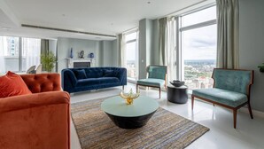 Un salon moderne avec un canapé bleu, un fauteuil orange et un fauteuil vert. Une table basse noire avec un pied doré est placée au centre de la pièce. La pièce est dotée de grandes fenêtres avec des rideaux, d’une cheminée et offre une vue sur la ville.