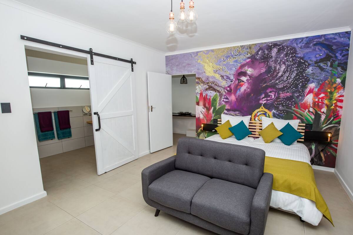 A modern bedroom with a grey sofa, a bed with colorful pillows, a mural on the wall, and a sliding door leading to another room.