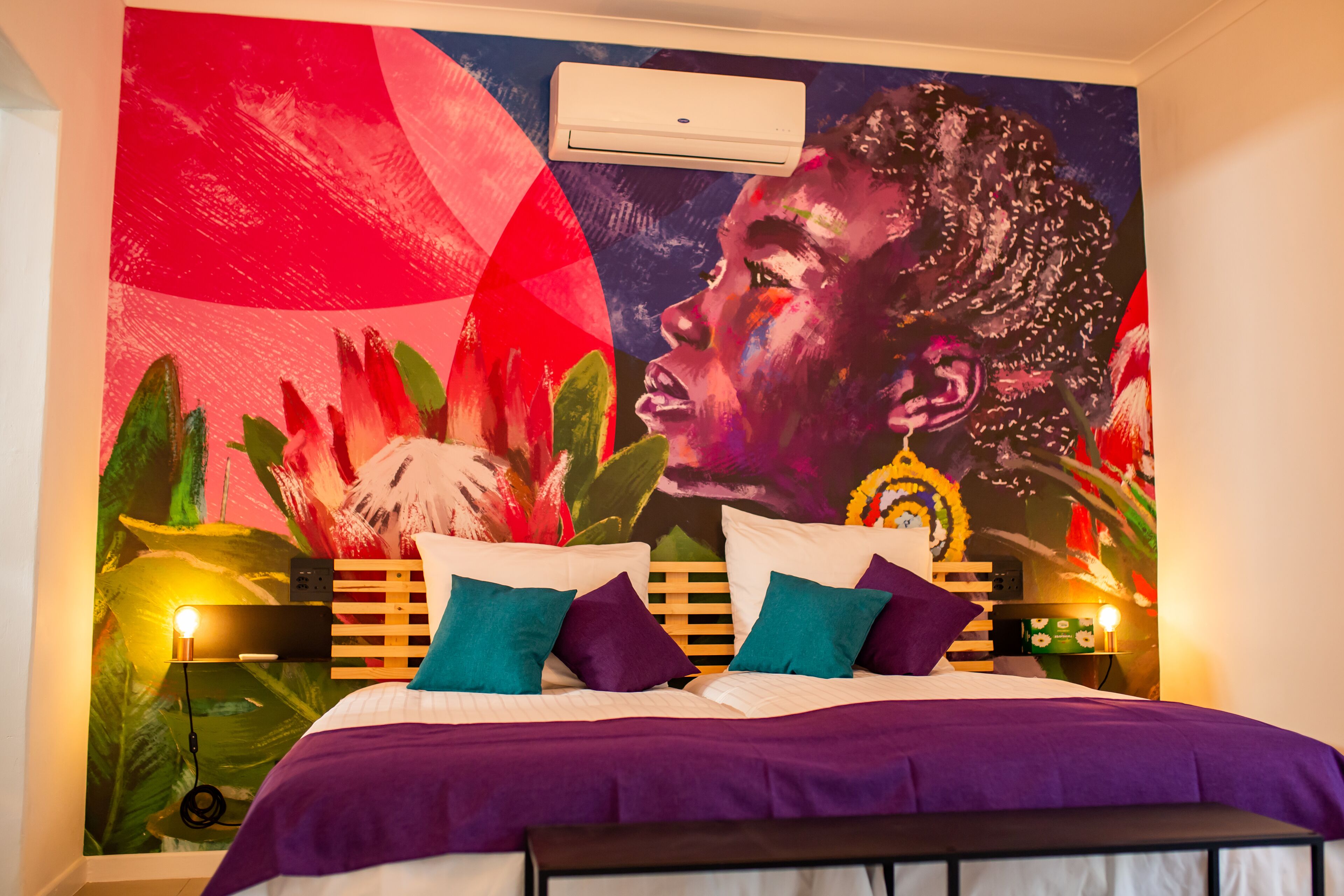 A bedroom with a large mural, a bed with purple bedding, and two bedside lamps.
