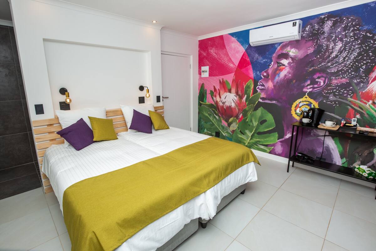 A modern bedroom with a large bed, colorful pillows, a vibrant mural, and a small side table with a coffee maker.