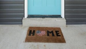 Eine türkisblaue Tür mit einem Kranz, einer Fußmatte mit dem Schriftzug „HOME“ und einer amerikanischen Flagge, sowie eine kleine Wandleuchte.