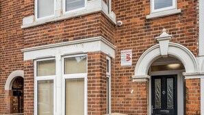 A two-story brick house with a black front door, white-framed windows, and a warning sign on the wall. - Studio Flat in Bedford Near Hospital & Stations (Bedford)