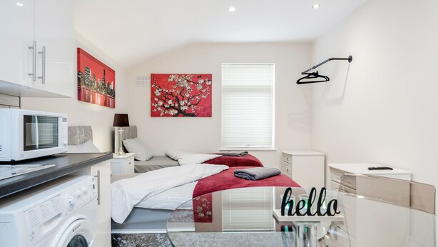 A modern bedroom with a bed, a washing machine, a glass table, and a cityscape painting. - Studio Flat in Bedford Near Hospital & Stations (Bedford)