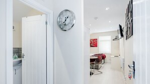 A modern kitchen with a white door, a wall-mounted clock, and a dining area with a red decorative piece. - Studio Flat in Bedford Near Hospital & Stations (Bedford)