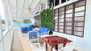 Un espace extérieur abrité, aménagé avec des chaises en plastique blanc et une table en bois, des coussins bleus et un mur vert.