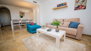 Un salon moderne avec un canapé beige, un fauteuil bleu, une table basse blanche et un coin repas comprenant une table blanche et des chaises grises.