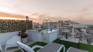Een dakterras met witte stoelen en een tafel, een plant in pot, en uitzicht op een historisch stadsbeeld bij zonsondergang.