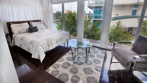 Ein Schlafzimmer mit einem großen Bett, einem Glastisch, einer Couch und Blick auf ein Gebäude und Bäume durch große Fenster.