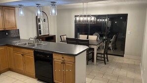 A modern kitchen with a central island, wooden cabinets, and a dishwasher. There is a dining area with a table and chairs, and a large window providing natural light. - Quite Waterfront Villa+Heated Pool+ Close to Dining+Shopping+Entertainment (Cape Coral)