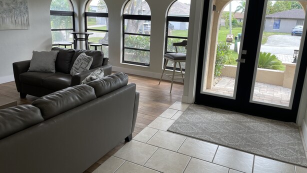 A living room with a sofa, a dining table, and chairs, large arched windows, and a door leading outside. - Quite Waterfront Villa+Heated Pool+ Close to Dining+Shopping+Entertainment (Cape Coral)