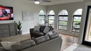 A modern living room with a grey sofa, a wooden coffee table, and a television displaying a show. There are large windows with arched tops, a dining area with a table and chairs, and a grey cabinet with drawers. - Quite Waterfront Villa+Heated Pool+ Close to Dining+Shopping+Entertainment (Cape Coral)
