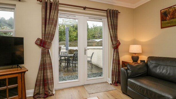 A living room with a TV, a leather sofa, a lamp, and a sliding glass door leading to an outdoor patio with a table and chairs. - Castle Crag (Keswick)