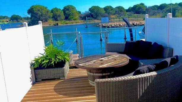 Eine Holzterrasse mit Gartenmöbeln, darunter ein Rattansofa und ein runder Tisch, mit Blick auf ein Gewässer, im Hintergrund Boote und Grünpflanzen.
