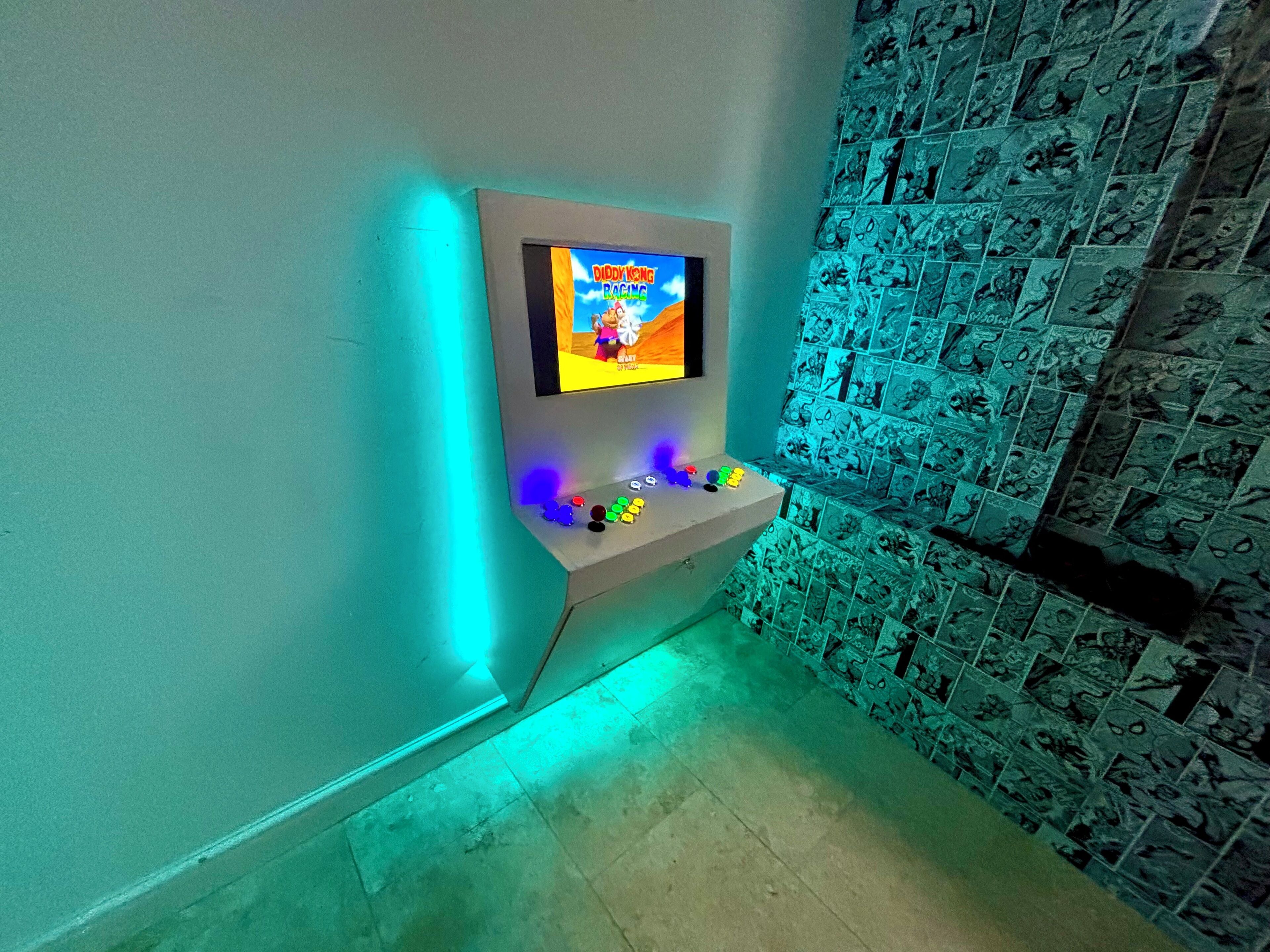 Ein Zimmer mit einem an der Wand befestigten Fernseher, der 'Diddy Kong Racing' zeigt, einem Regal mit bunten Gegenständen und einer Wand, die mit comicartigen Fliesen verziert ist.
