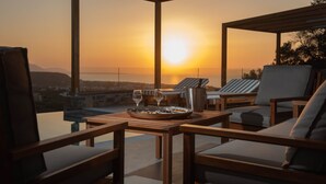 Eine Terrasse mit Holzmöbeln, einem Tisch mit Speisen und Gläsern sowie Blick auf den Sonnenuntergang über der Stadt.