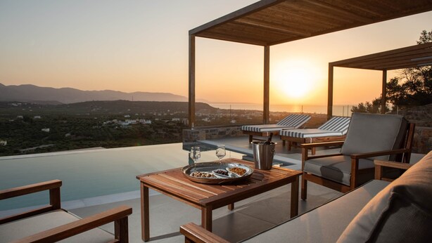 Eine Terrasse mit Holztisch, Liegestühlen und Blick auf den Sonnenuntergang über den Bergen.