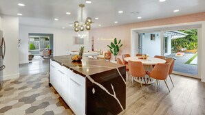A modern kitchen with a marble island, dining area with orange chairs, and a view of a garden through large windows. - ~NEW~Casa Miami~Luxury Oasis w/ Pool&Game Paradise (Miami)