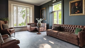 Un salon spacieux avec un canapé en cuir marron, un fauteuil assorti et une petite table d’appoint sur laquelle est posé un vase. De grandes fenêtres laissent entrer la lumière naturelle et offrent une vue sur la verdure environnante.
