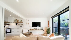 A modern living room with a beige sofa, a flat-screen TV mounted on the wall, and a large window offering a view of greenery.