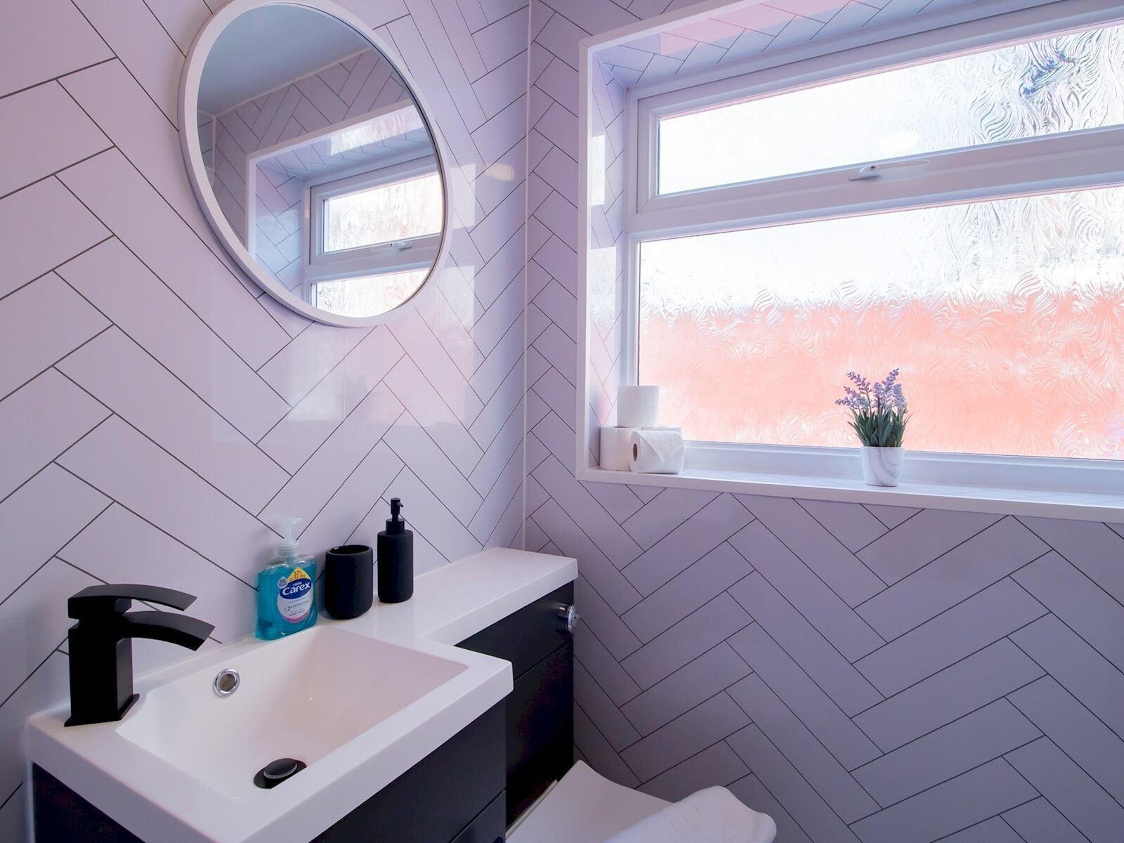 A bathroom with a white sink, black faucet, and a mirror above it. There is a window with a view of a pink wall and a plant on the windowsill.