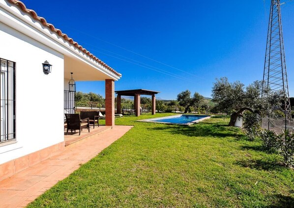 A white building with a tiled patio, a swimming pool, and a pergola.