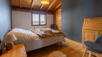Una camera da letto con soffitto in legno, un letto con copripiumino bianco, una lampada su un comodino in legno, una sedia e una vista dalla finestra.