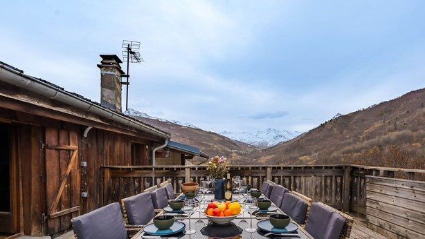 A wooden deck with a dining table set for six, surrounded by wicker chairs, with a mountain backdrop. - Chalet “Le petit Paradis” in the heart of the 3 valleys! (Les Belleville)